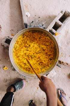 Top-down shot of a hand stirring yellow rice in a large pot outdoors.