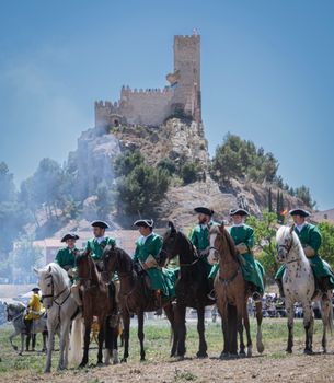 Men in traditional clothing on horseback in front of Almansa Castle during a historical reenactment.