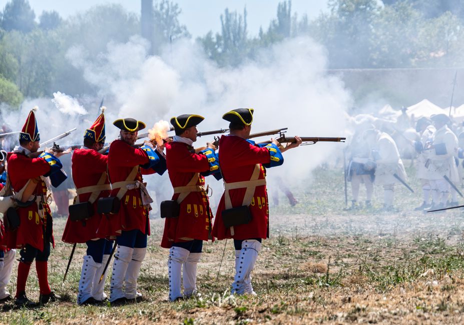 Reenactors in vibrant uniforms firing muskets during a historical battle event.
