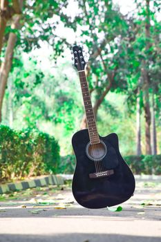 Black acoustic guitar standing outdoors in a sunny, vibrant park setting.