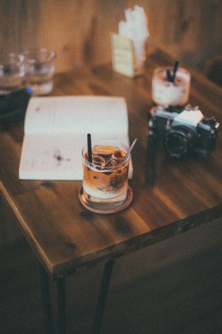 A warm, inviting scene with iced coffee, a camera, and an open book on a wooden table indoors.