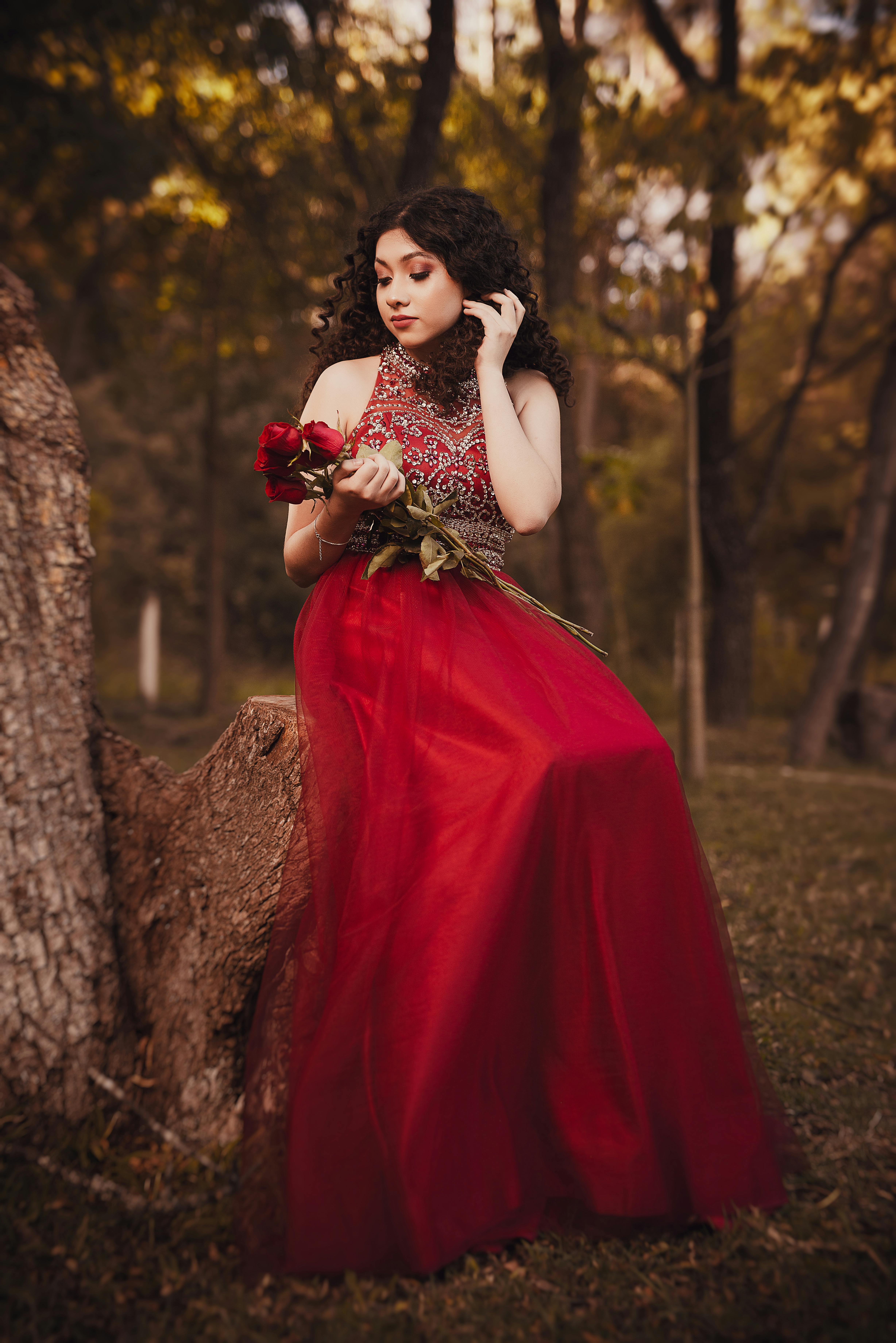 A brunette woman in an elegant red dress holds roses in a woodland scene, exuding grace and style.