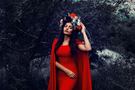 Beautiful woman in a red dress and flower crown posing in a mystical forest setting.