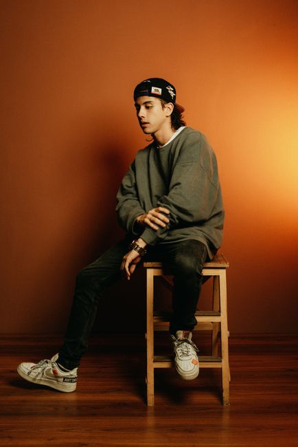 Teenage boy in fashion attire seated on a stool against warm background.