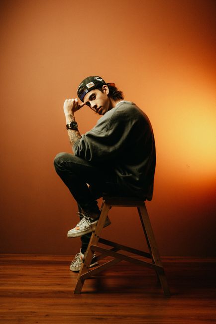 Portrait of a young man in casual fashion seated on a wooden stool in a studio setting.
