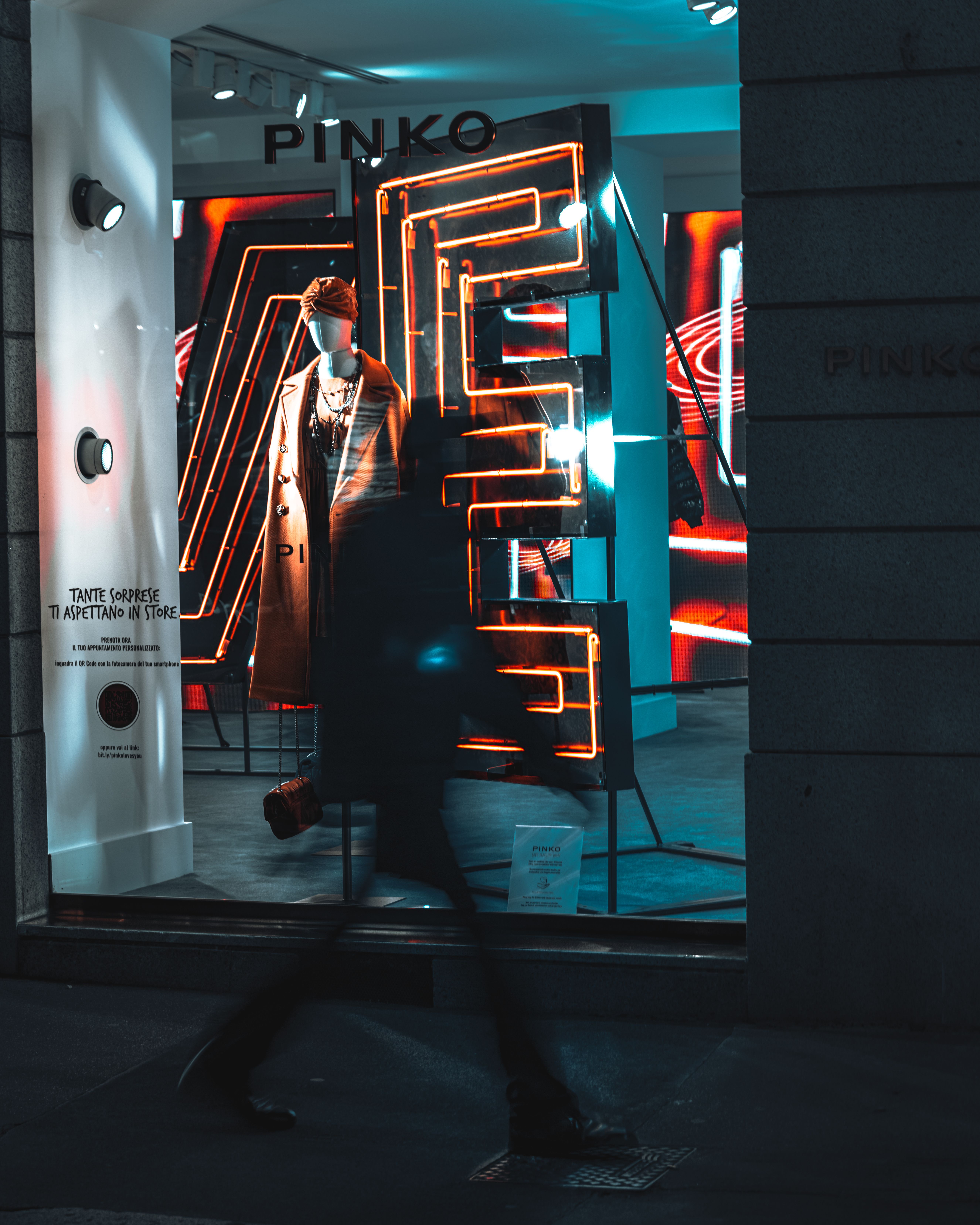 A blurred pedestrian passes a fashion storefront featuring neon lights and a mannequin in stylish clothing.