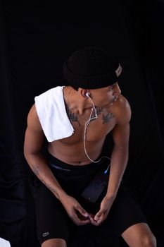 Portrait of a young man in sportswear with tattoos, wearing earphones and a beanie, against a black background.