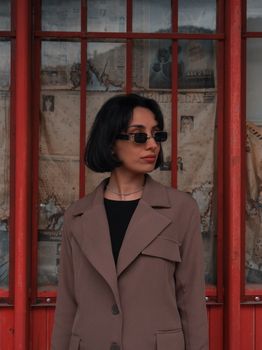 Stylish woman in sunglasses poses against a red urban backdrop, exuding confidence.