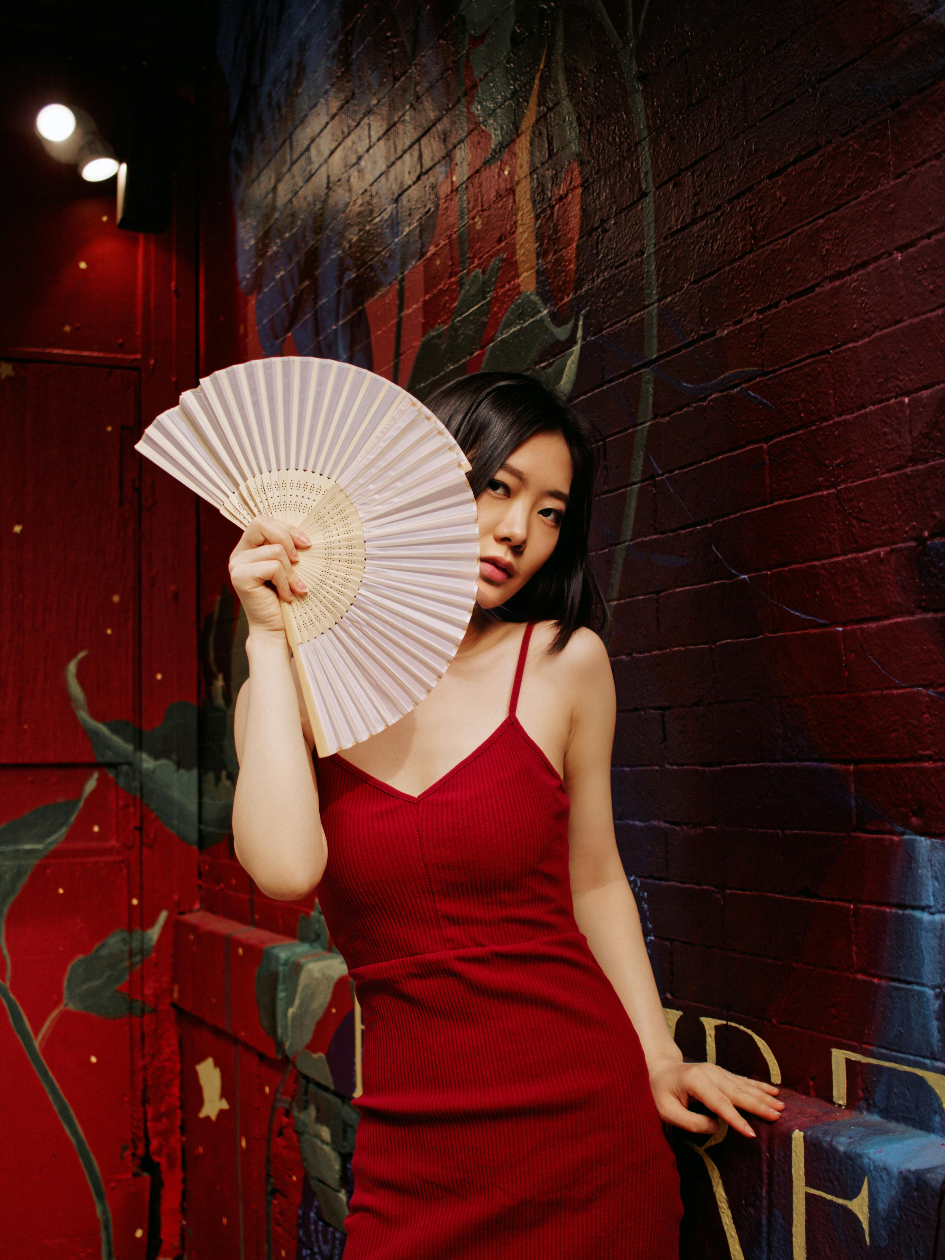 Stylish woman in a red dress poses elegantly with a fan against a vibrant wall.