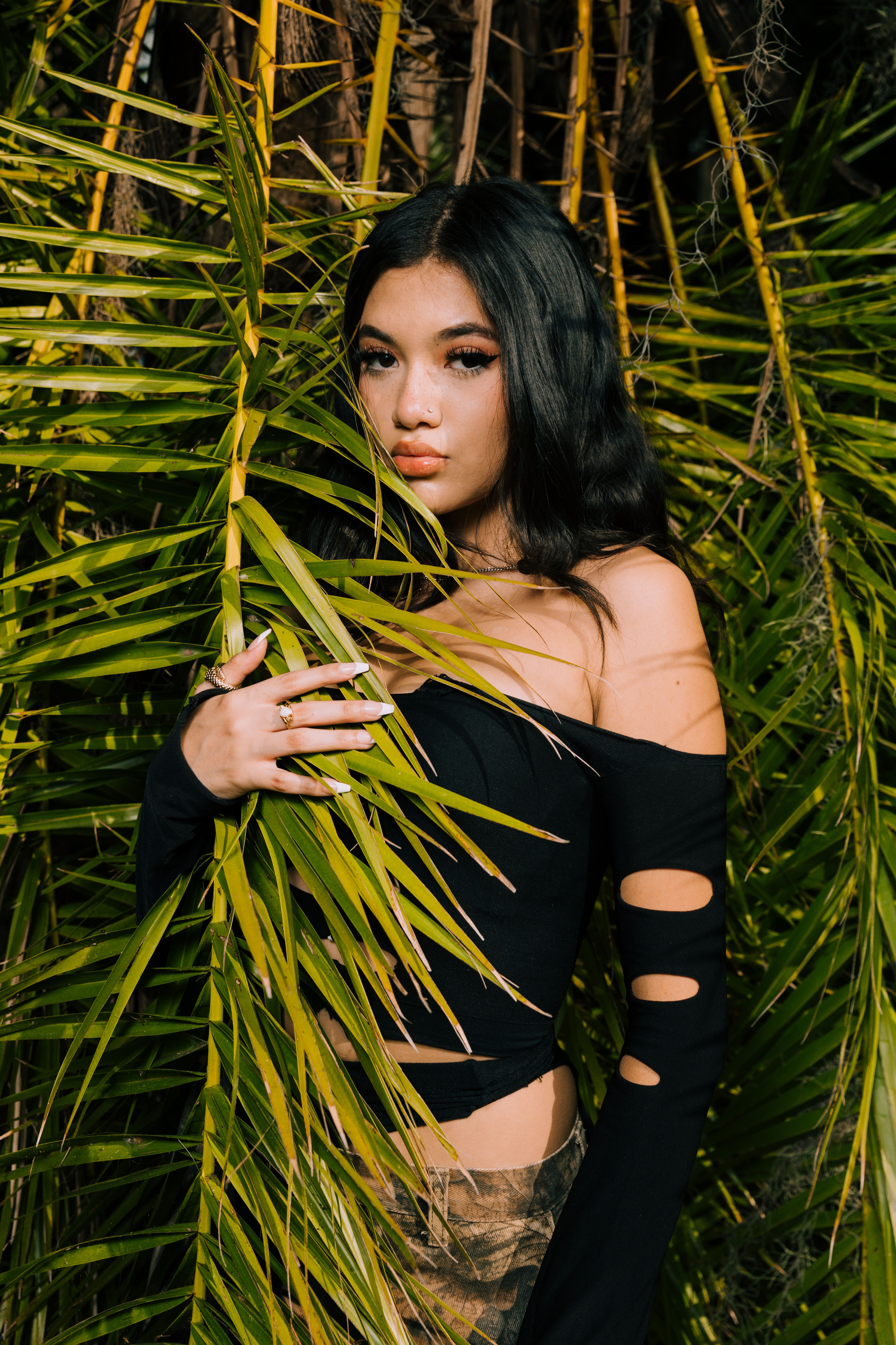 Young woman in black blouse posing gracefully amidst vibrant green leaves.