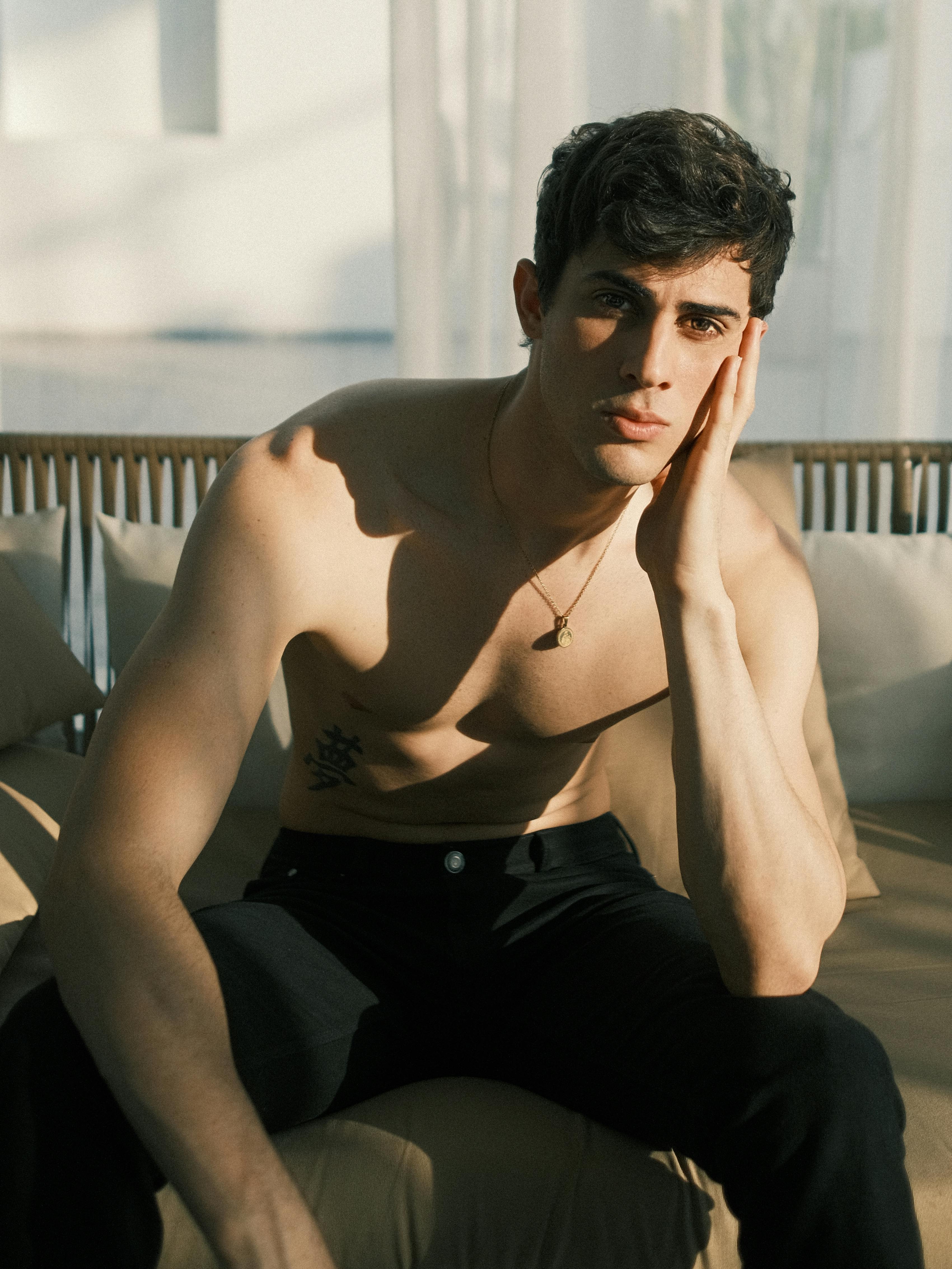 Free A topless man sitting with hand on cheek, bathed in soft indoor light. Ideal for lifestyle and fashion themes. Stock Photo