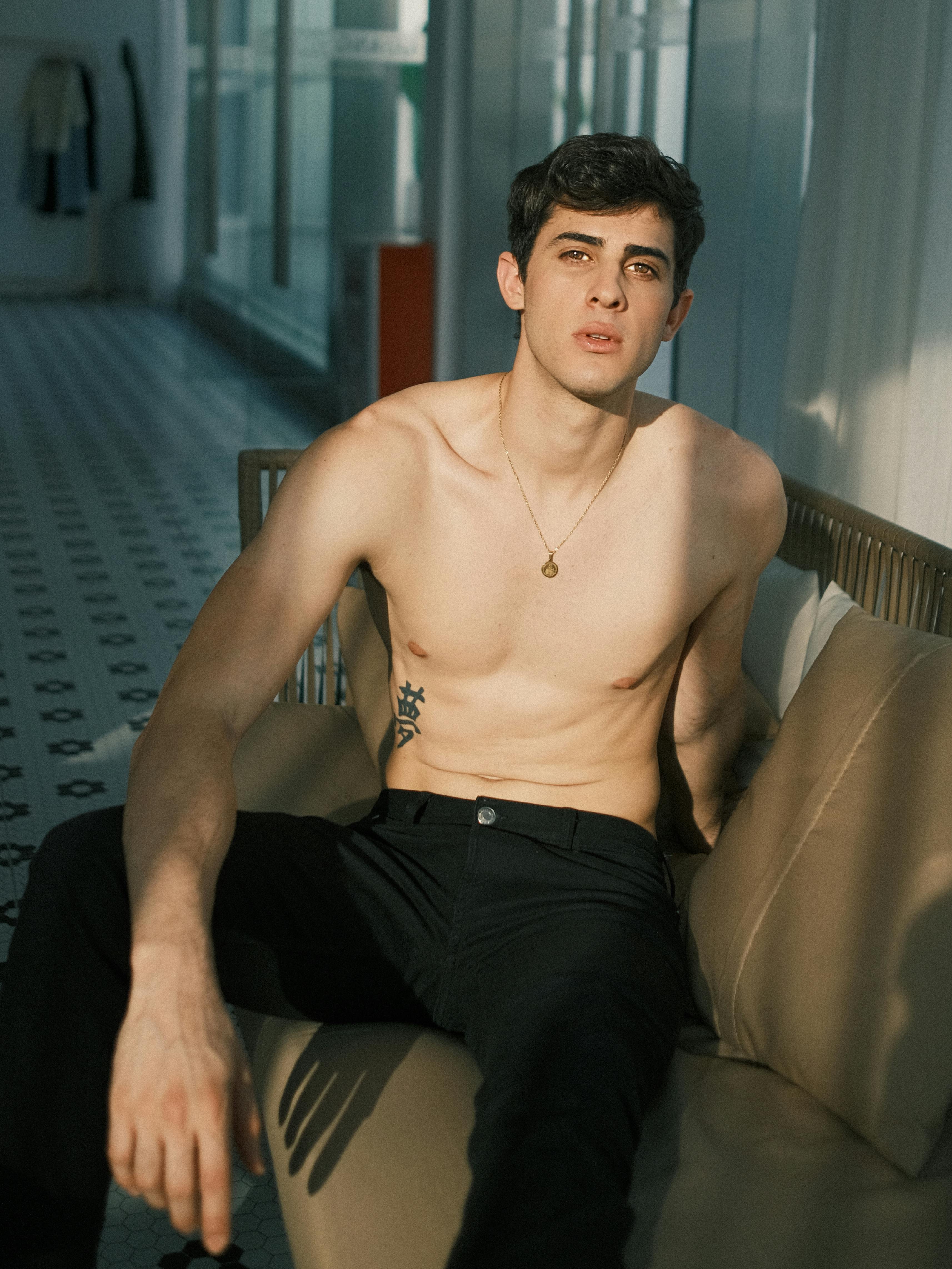 A young man posing topless on a couch in natural indoor lighting.