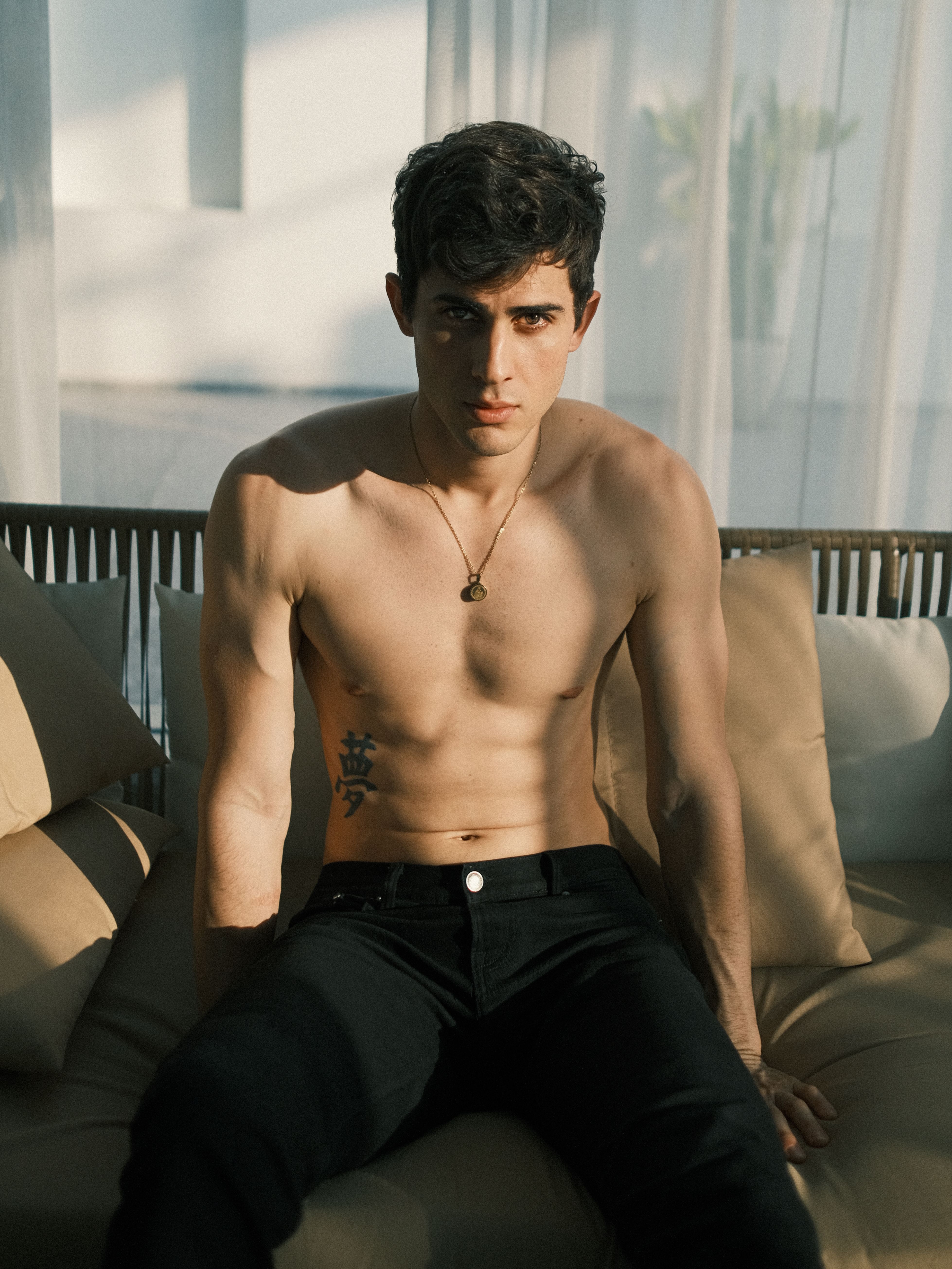 Shirtless young man posing on a sofa in a modern indoor setting with natural light.