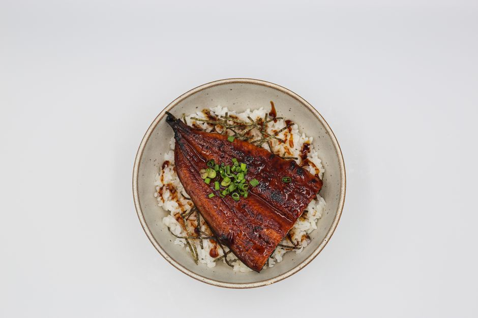 Glazed unagi fillets with dark caramelized kabayaki sauce laid over a bowl of steamed rice, garnished with strips of nori and a sprinkle of sansho pepper