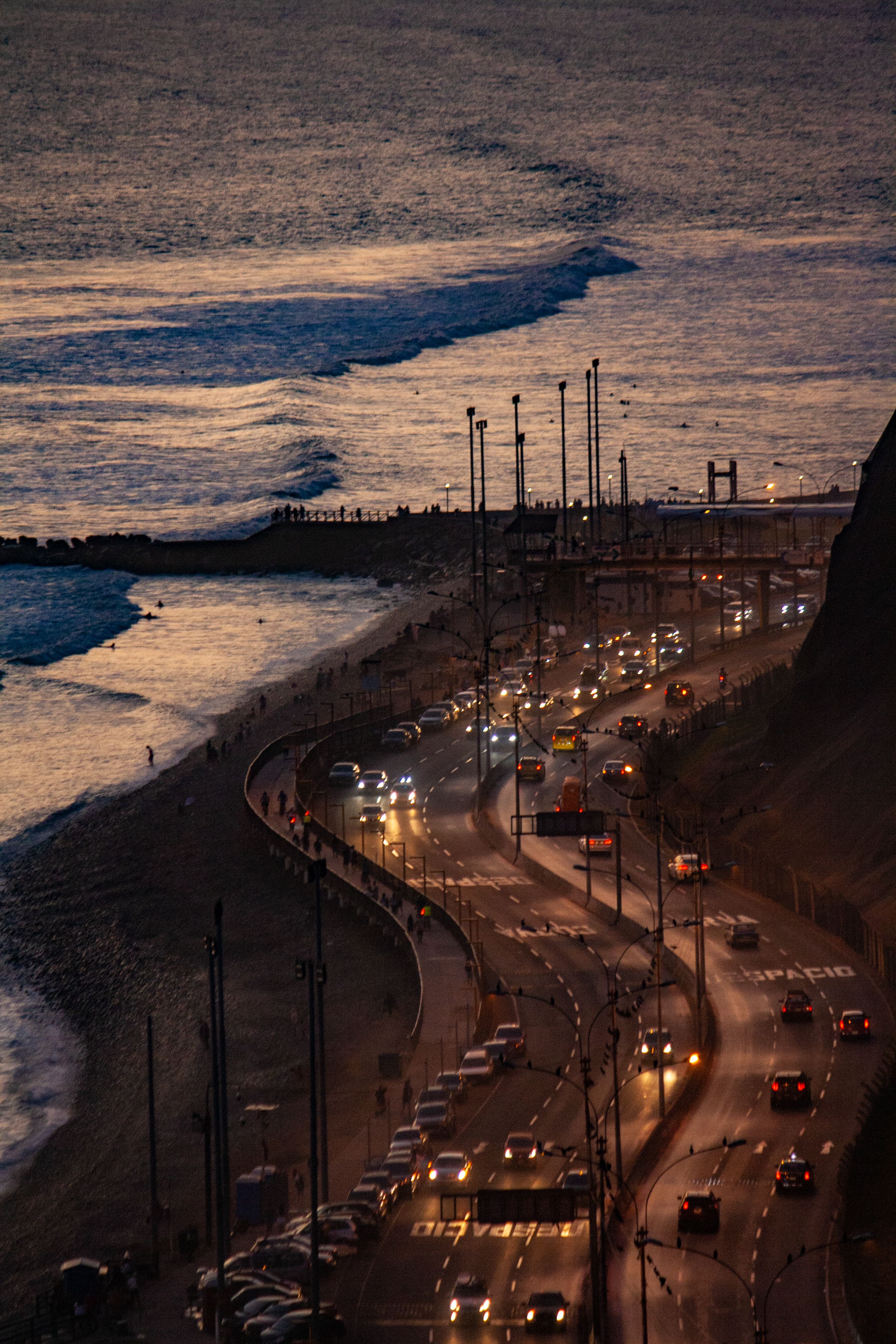 Coastal road beside the ocean at dusk with glowing car lights.