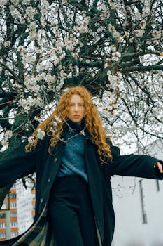 Redheaded woman in a coat standing under a blooming tree in Kyiv, ideal for fashion photography.