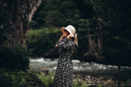 Free A woman in a floral dress and hat stands beside a river in a lush forest setting in Türkiye. Stock Photo