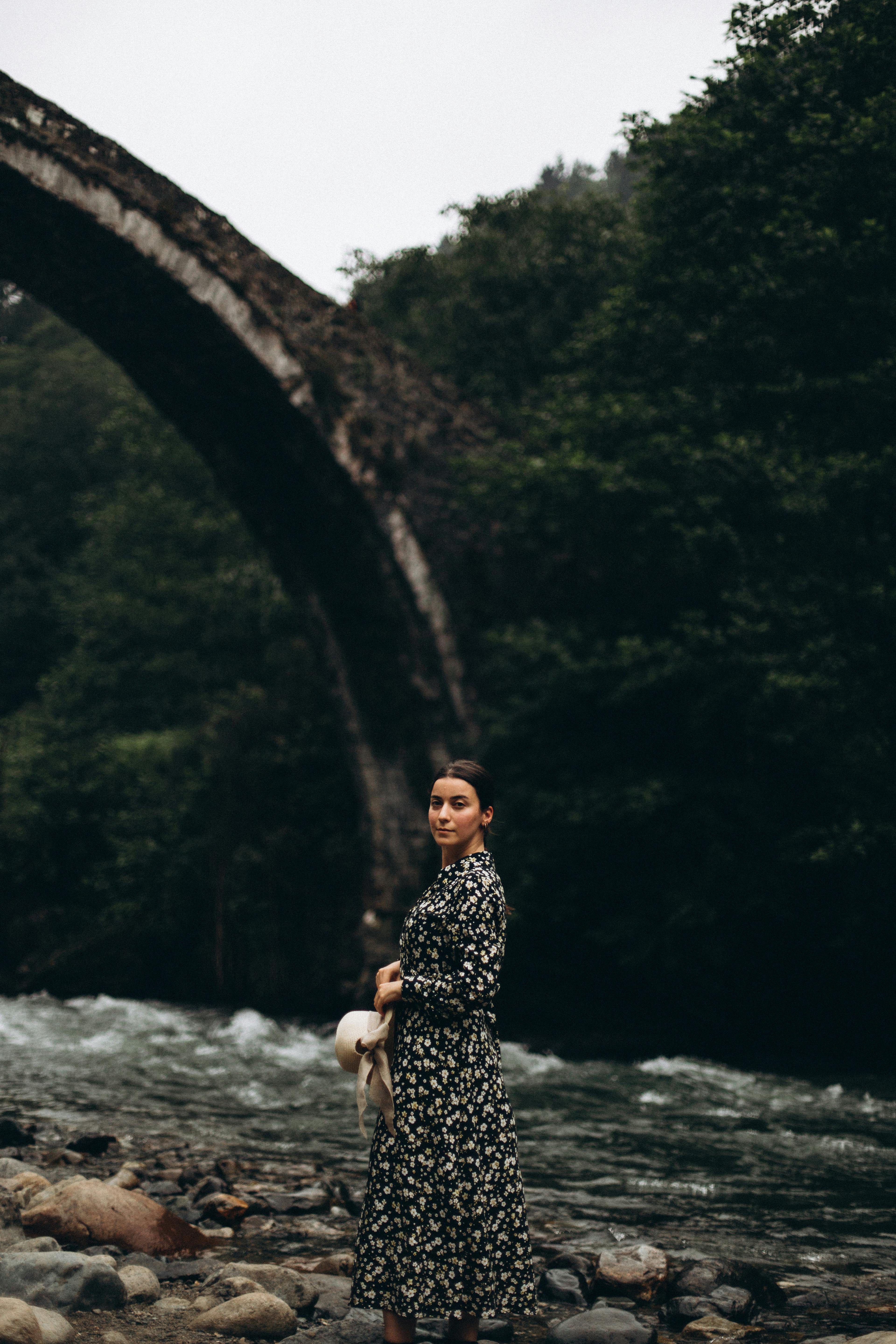 A woman stands by a scenic river under a historic stone bridge in Türkiye, wearing a floral dress.