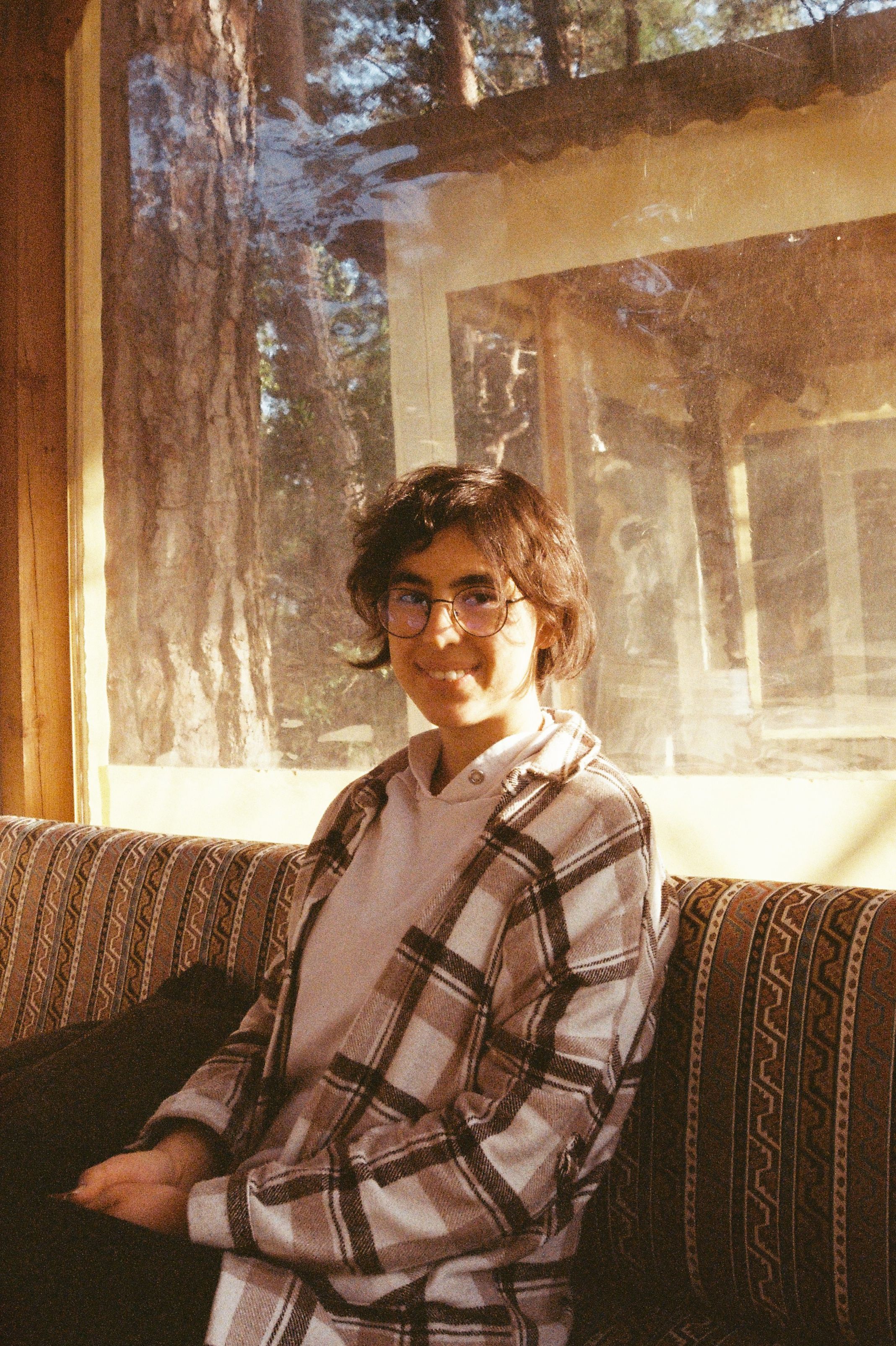 A woman in glasses smiles warmly indoors with natural light filtering through a window.