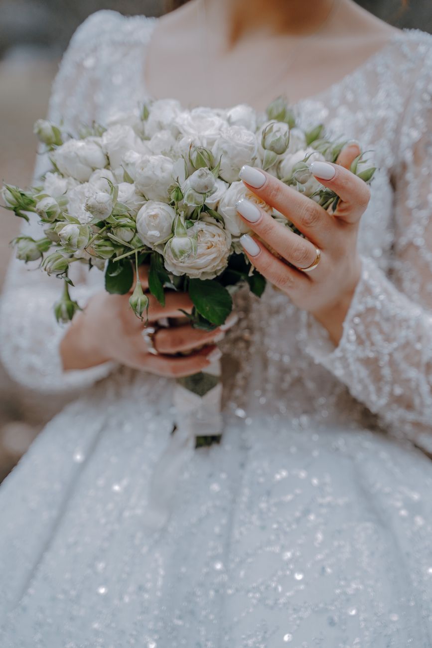 A close-up of an elegant bride holding a white rose bouquet, showcasing wedding nails and dress.