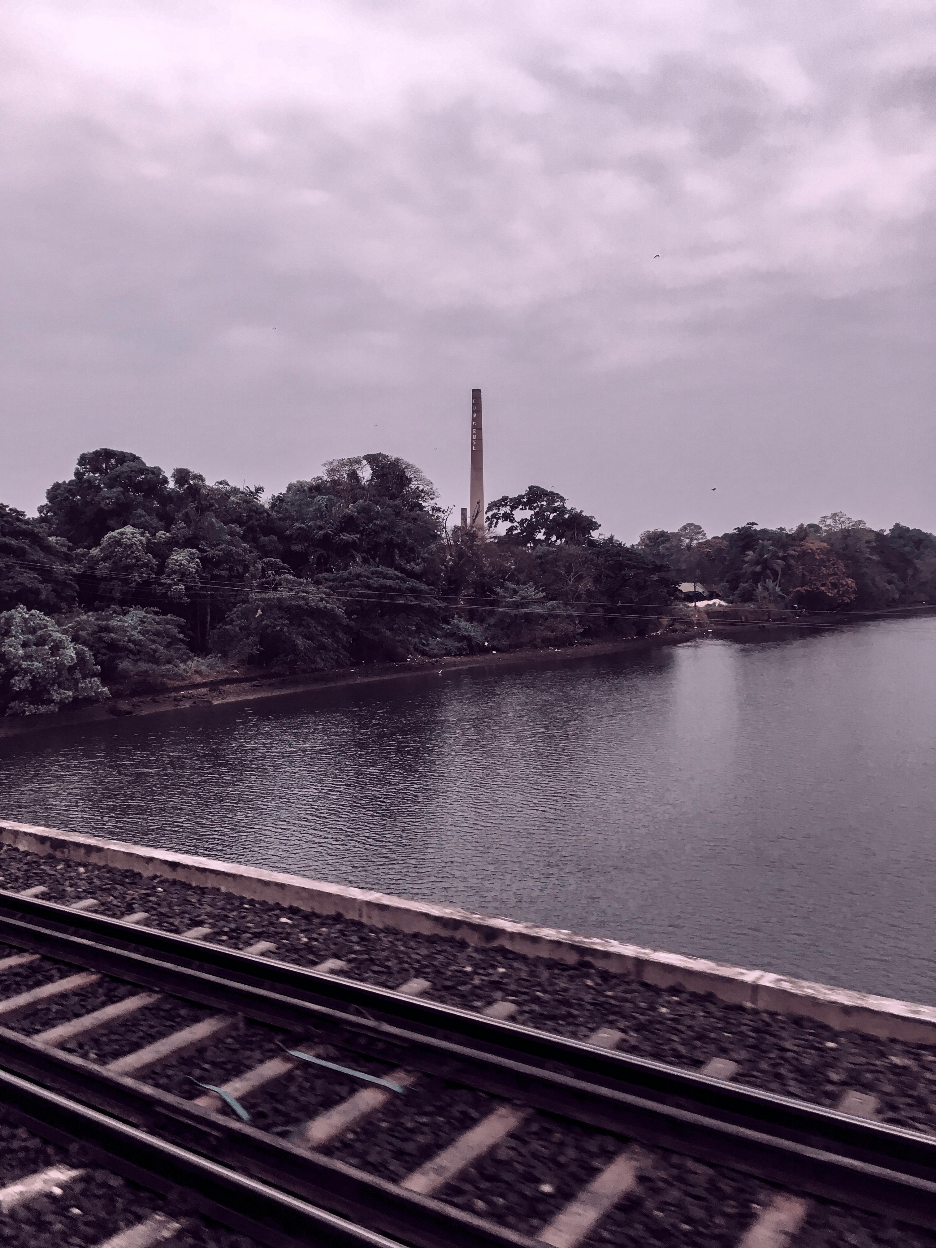 在多云的天空下，平静的河边景色中可见火车轨道与郁郁葱葱的绿植