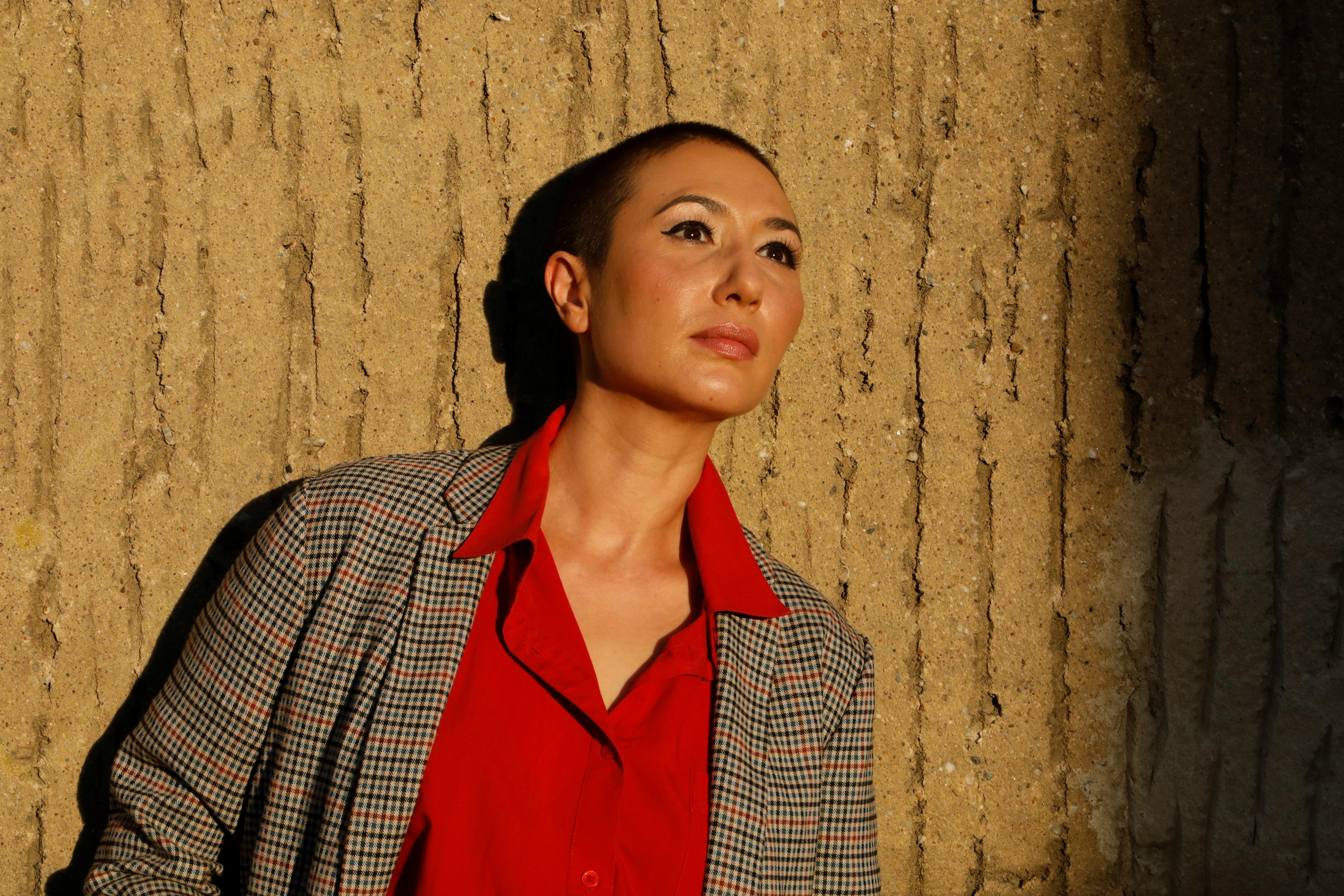 Elegant woman in red shirt posing against textured wall in warm sunlight.