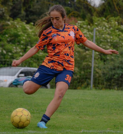 Free Young woman in orange soccer jersey kicking ball during outdoor match on grassy field. Stock Photo