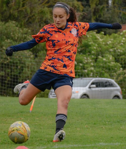 Free Woman in orange jersey playing soccer, mid-kick on a grassy field. Stock Photo