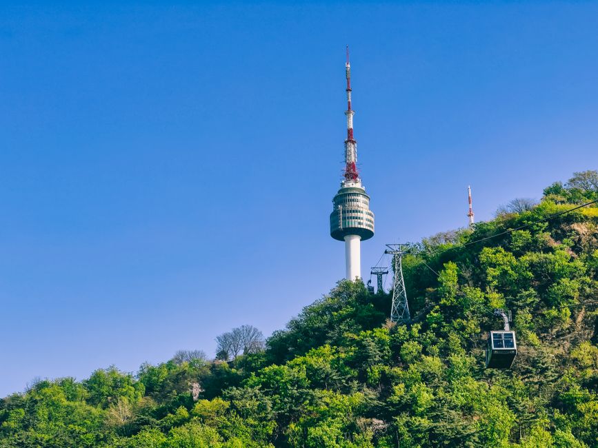 View of Namsan Tower with a moving cable car in Seoul, surrounded by lush greenery under a clear blue sky.