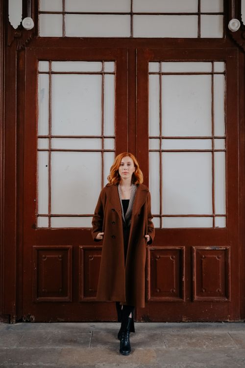 Free Fashionable woman with red hair in a brown coat stands confidently by large wooden doors, showcasing urban style. Stock Photo