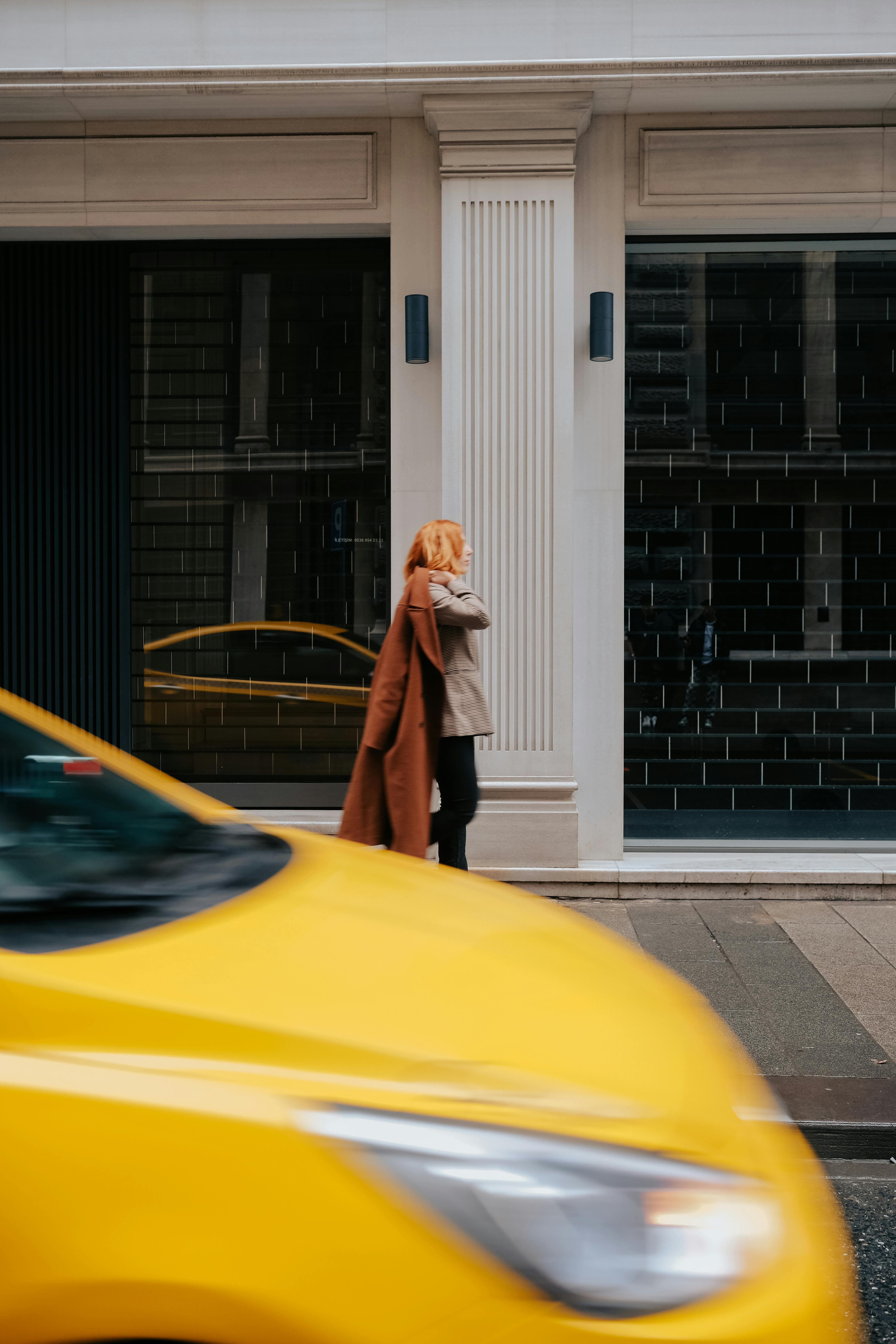 A woman adjusts her coat while a yellow taxi speeds by in the city, creating a dynamic urban scene.