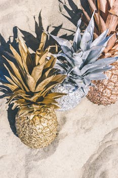 Artistic arrangement of gold, silver, and bronze pineapples on sand representing summer vibes.