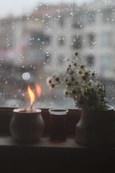 A serene scene of a candle and chamomile flowers by a window during a rainy day, creating a warm and peaceful ambiance.