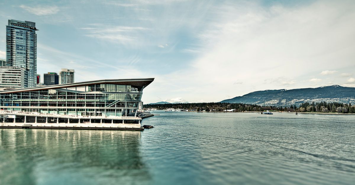 A stunning view of a modern waterfront building with mountains and urban architecture under a clear blue sky.