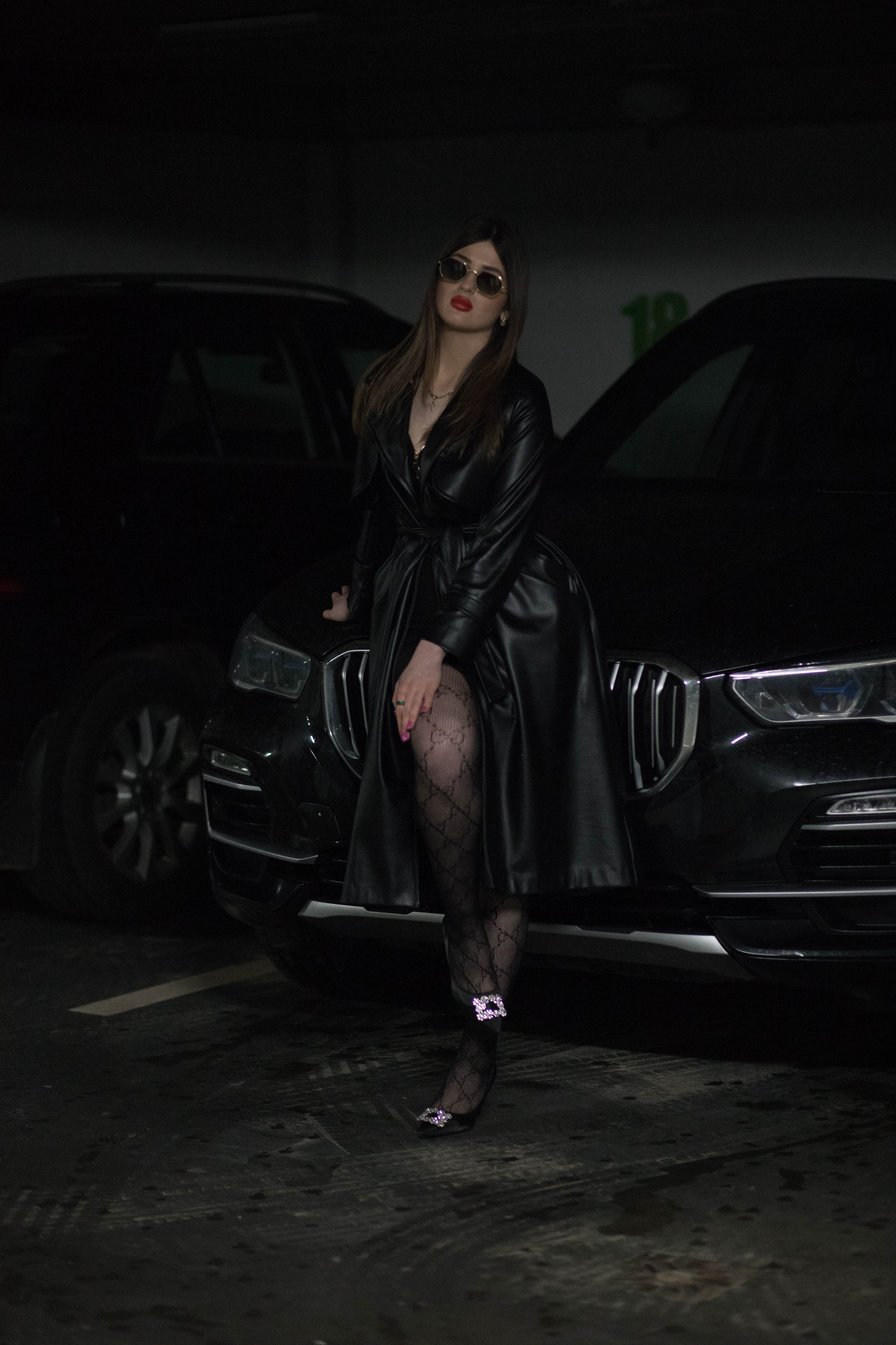 Stylish woman poses with a luxury car in a dimly lit parking garage.