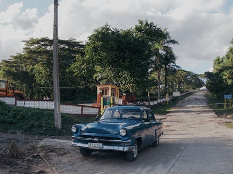 A retro blue car parked on a rural road surrounded by lush greenery under a clear sky.