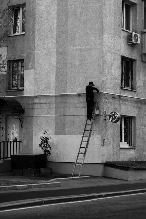 Black and White Photo of a Man Standing on a Ladder and Fixing a Building