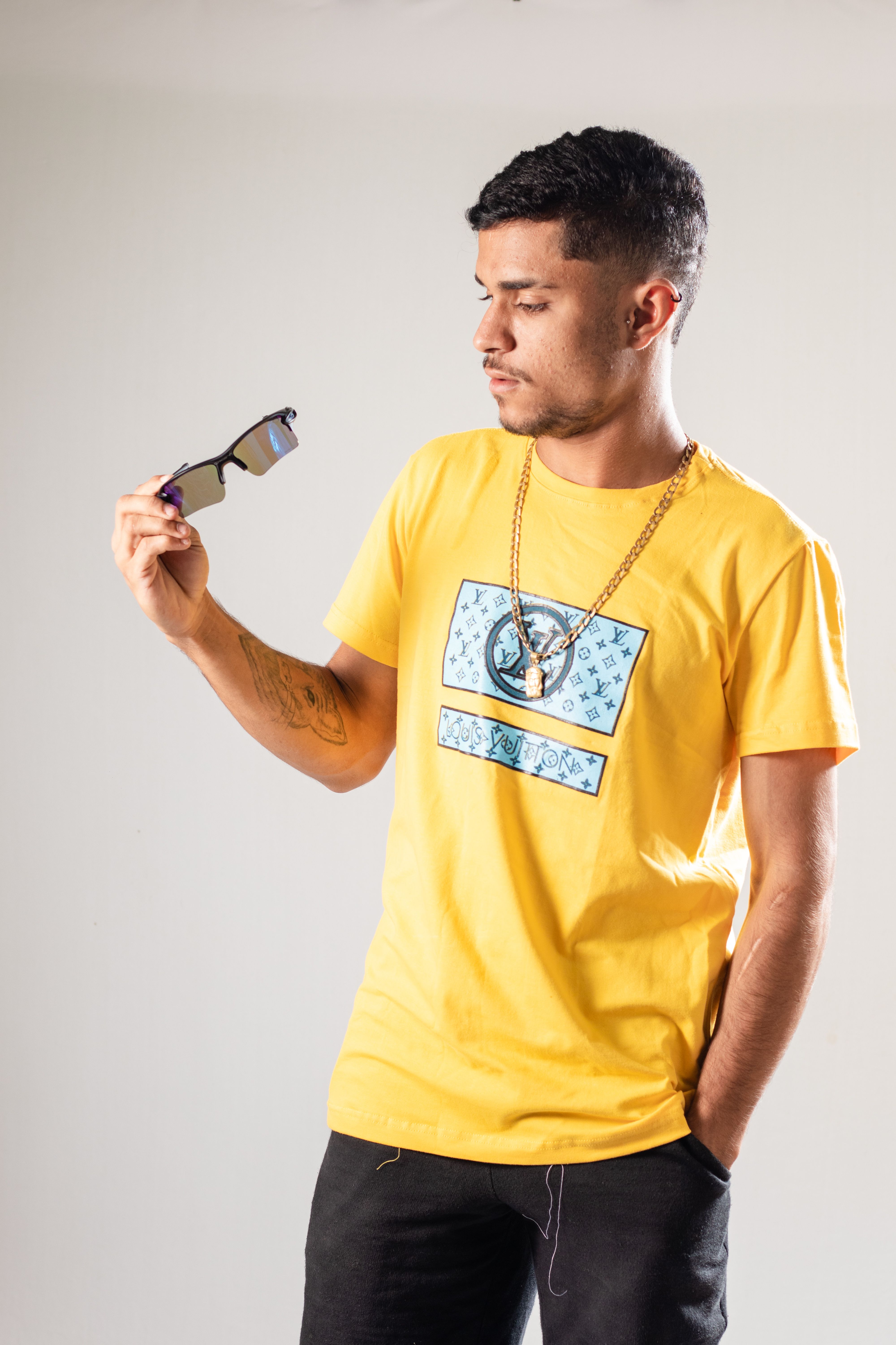 Stylish man in a yellow t-shirt with a necklace examining sunglasses in a studio setting.