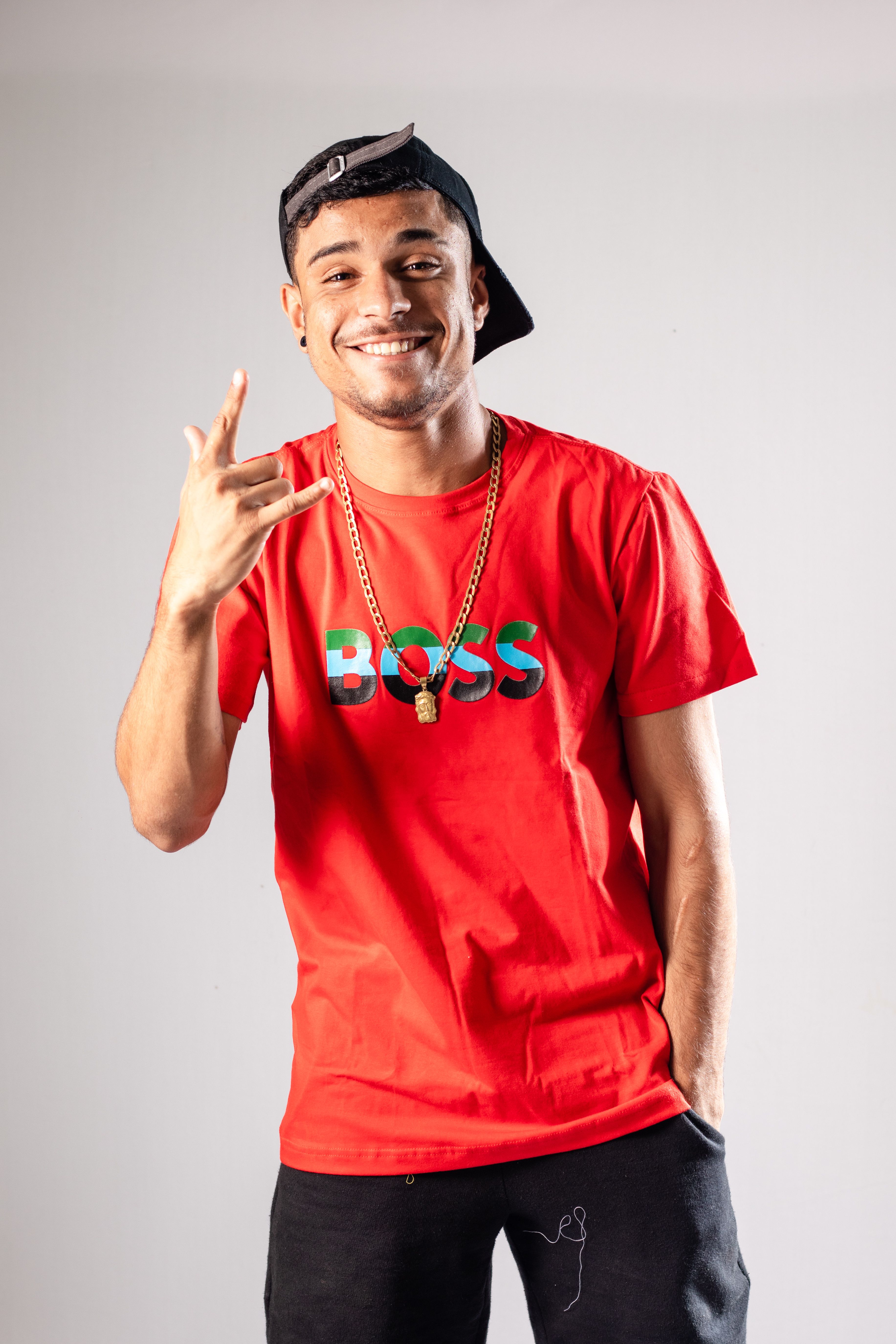 Young man wearing a red t-shirt and gold necklace, smiling and gesturing in a studio shoot.