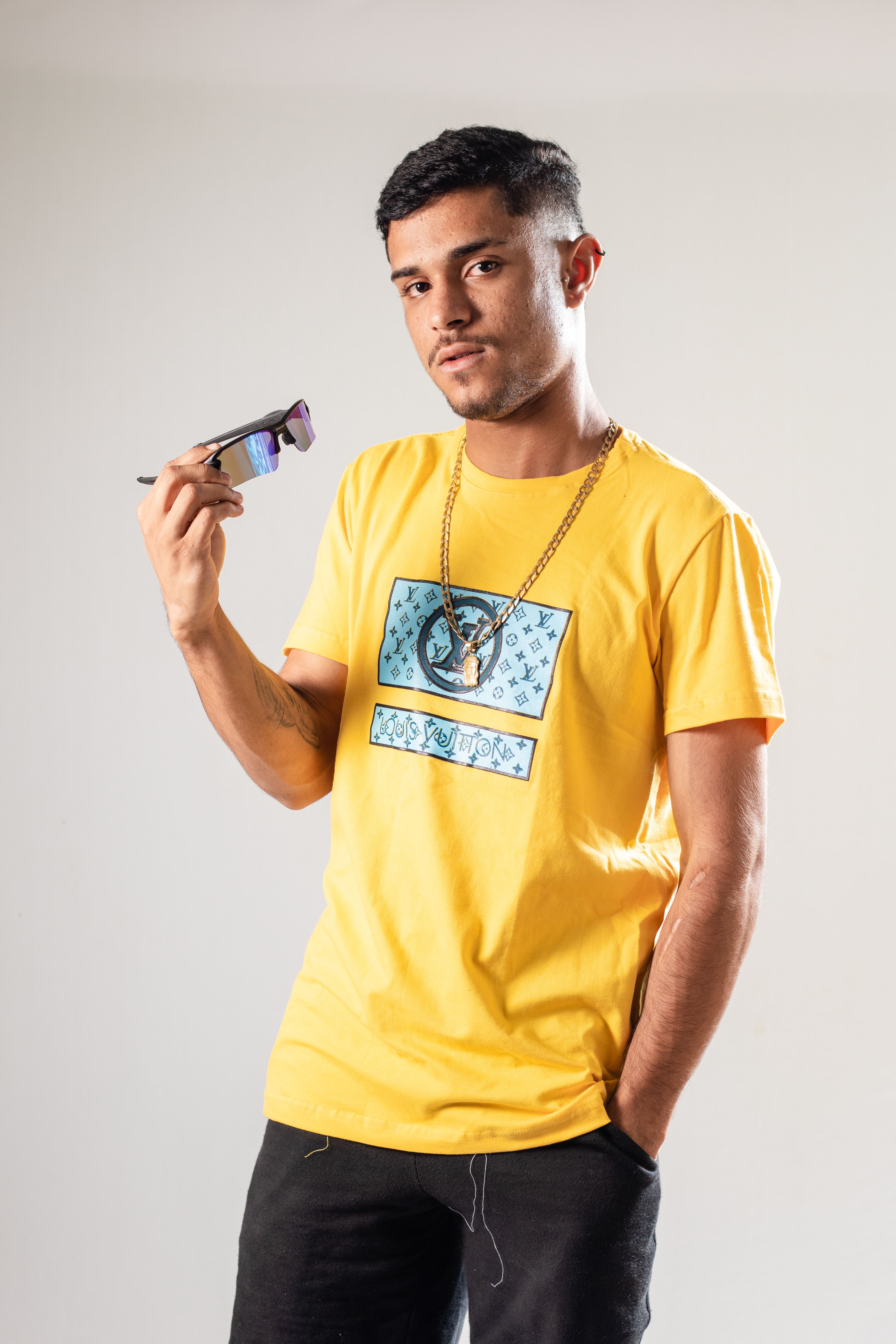 Fashionable young man posing with sunglasses in a studio, wearing a yellow t-shirt and necklace.