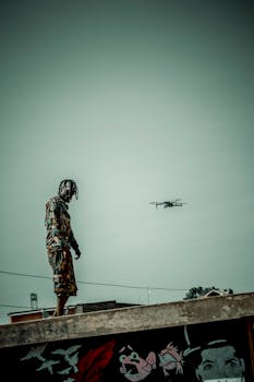 Camouflaged soldier stands on a rooftop with a drone in the sky, creating a dramatic silhouette.