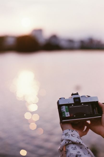 Close-up of hands holding an analog camera by a lake at sunset, with reflections and bokeh effects.