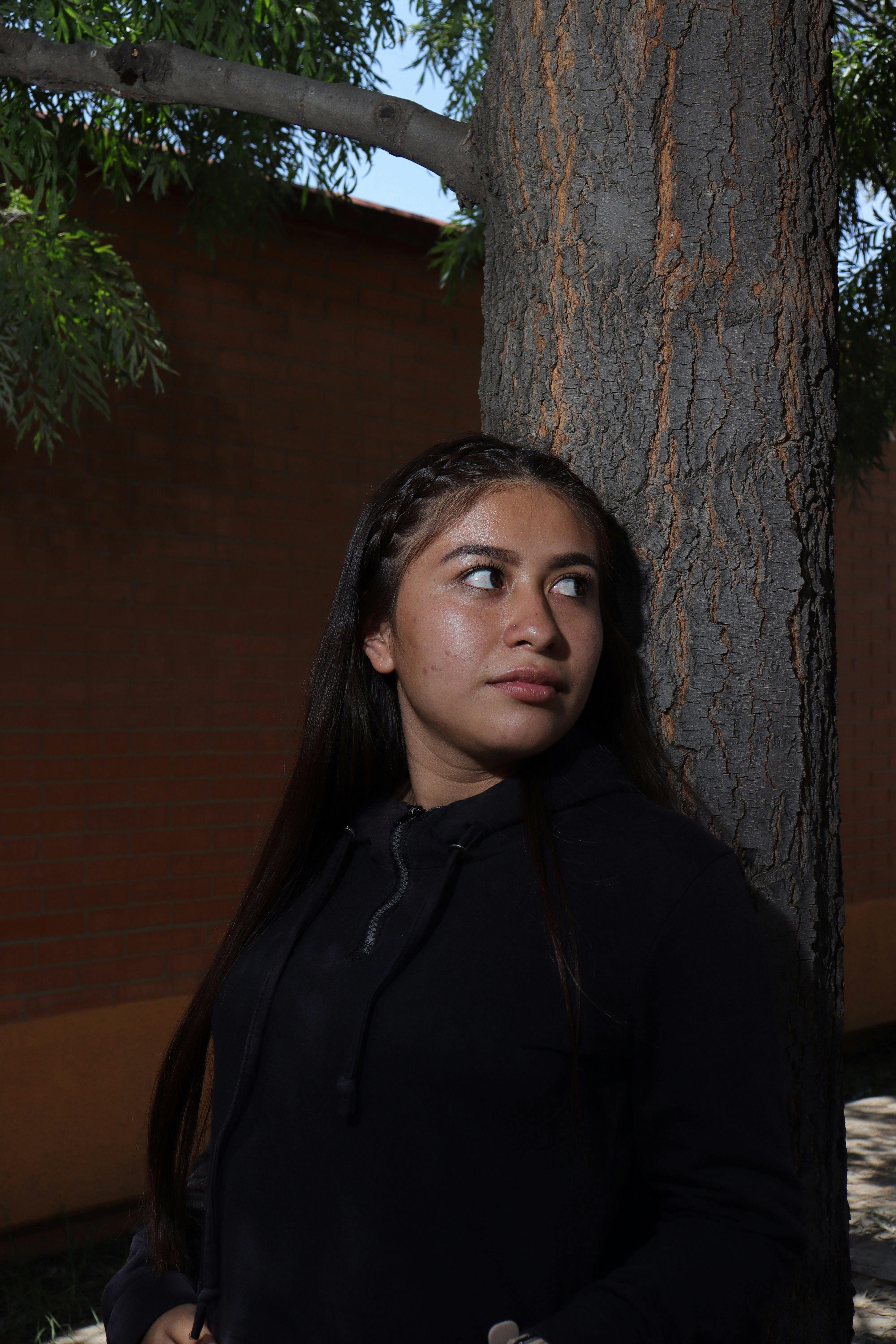 A young woman stands by a tree, looking away, dressed casually in a hoodie in an outdoor setting.