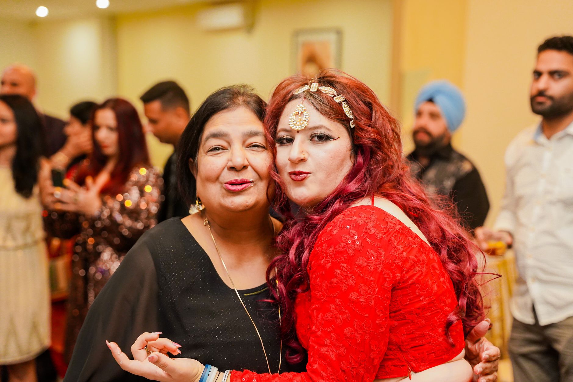 Women enjoying a joyful moment during an Indian wedding reception with guests in the background.