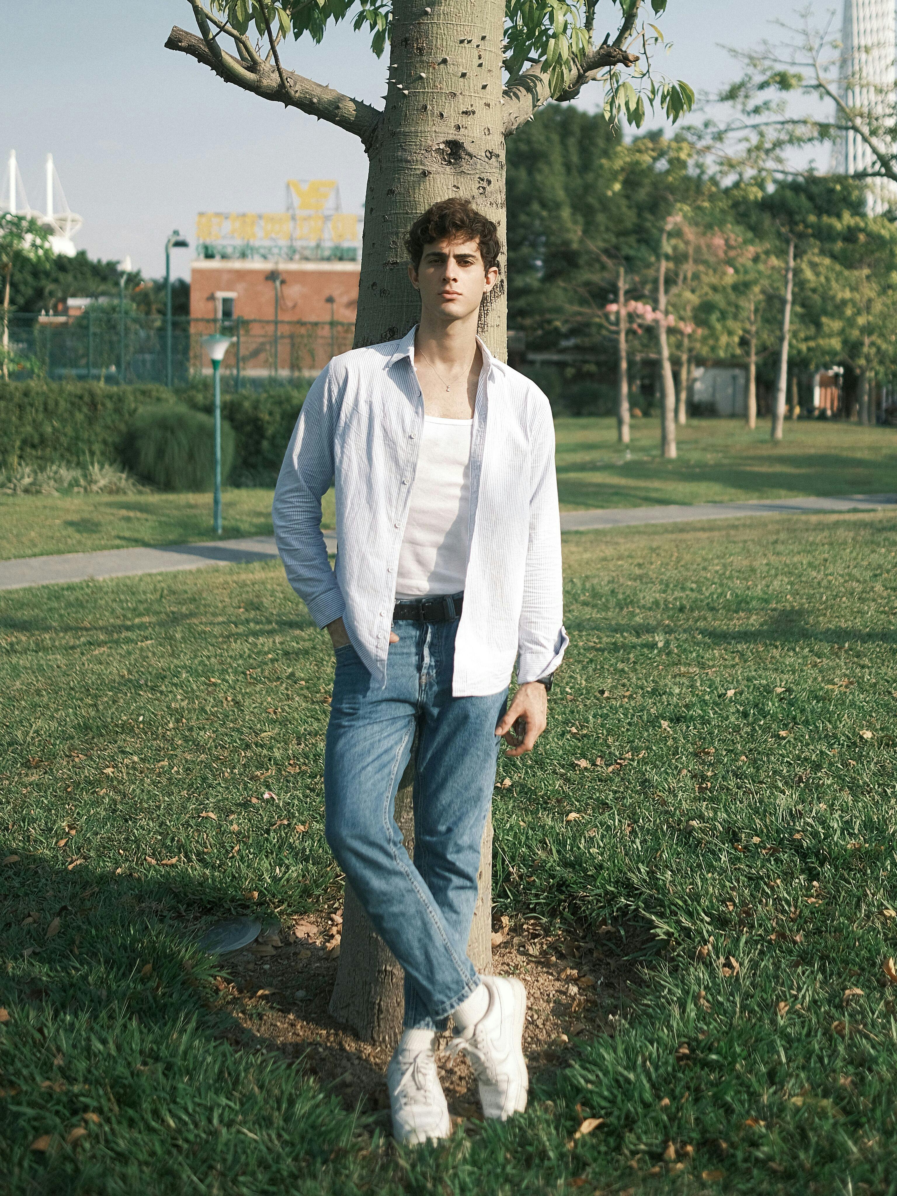 A young man in a casual white shirt and denim jeans leans against a tree in a park setting.