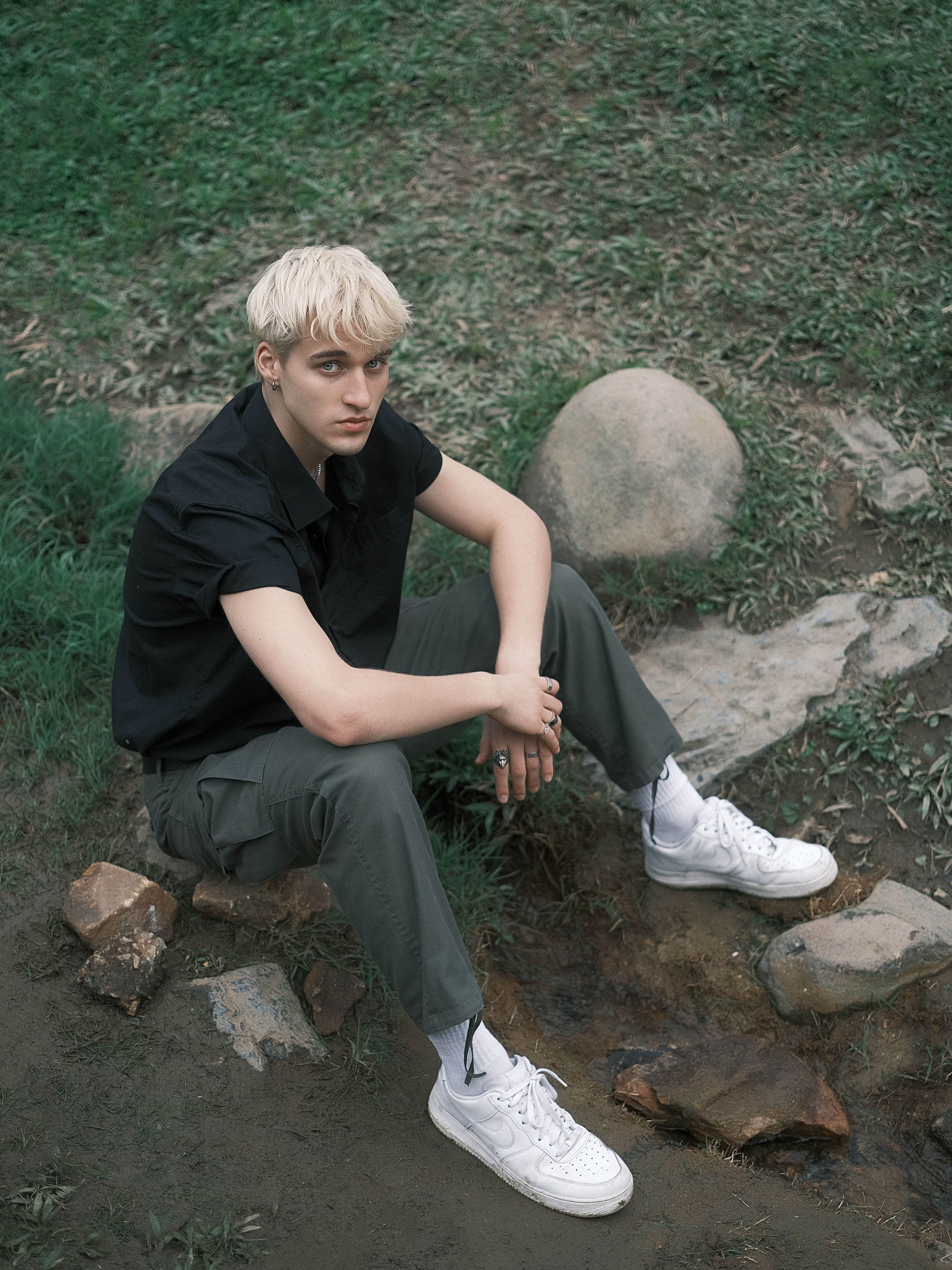 Young man with blond hair sitting on grass outdoors in casual streetwear, looking thoughtful.