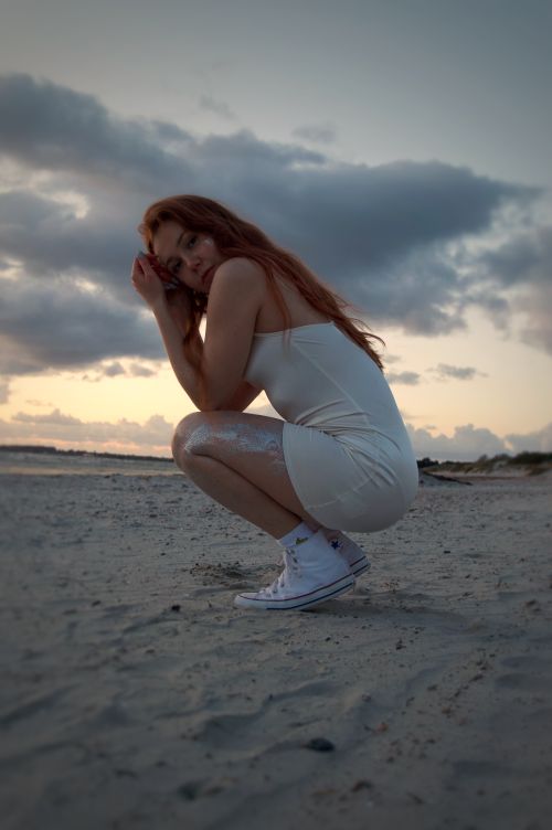 Free A young woman with long red hair crouching on a sandy beach during sunset. Stock Photo