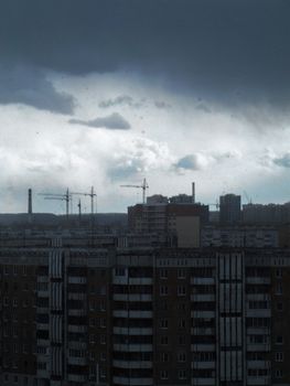 Dark clouds loom over a modern cityscape, highlighting tall buildings and construction cranes.
