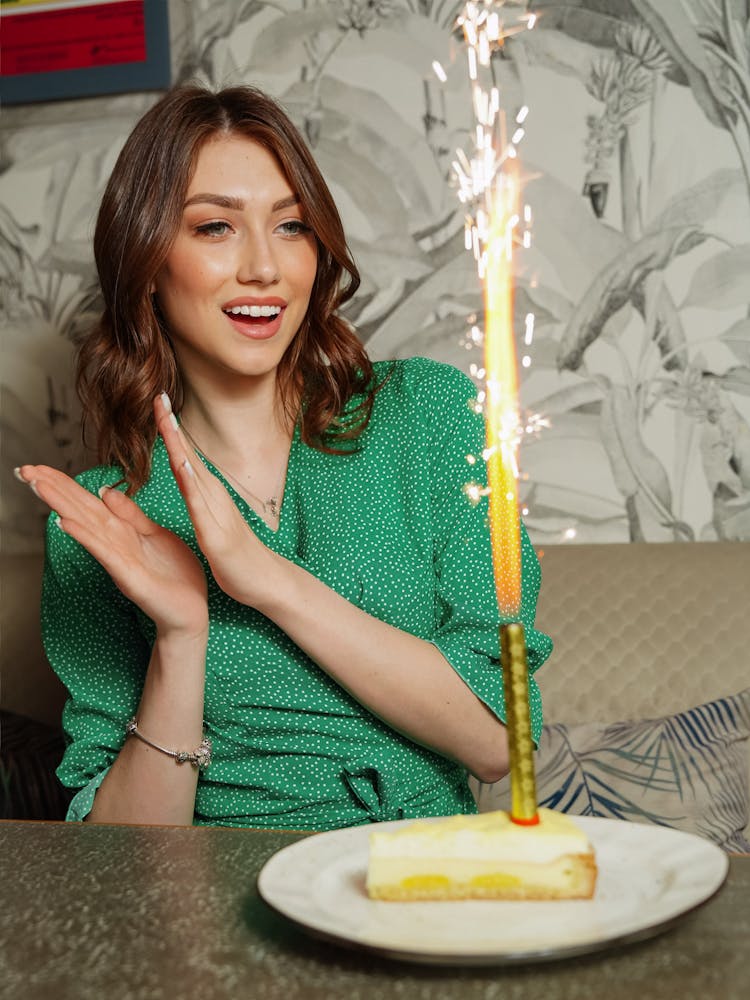 Young Woman Sitting At The Table With A Candle In A Slice Of Cake In Front Of Her 