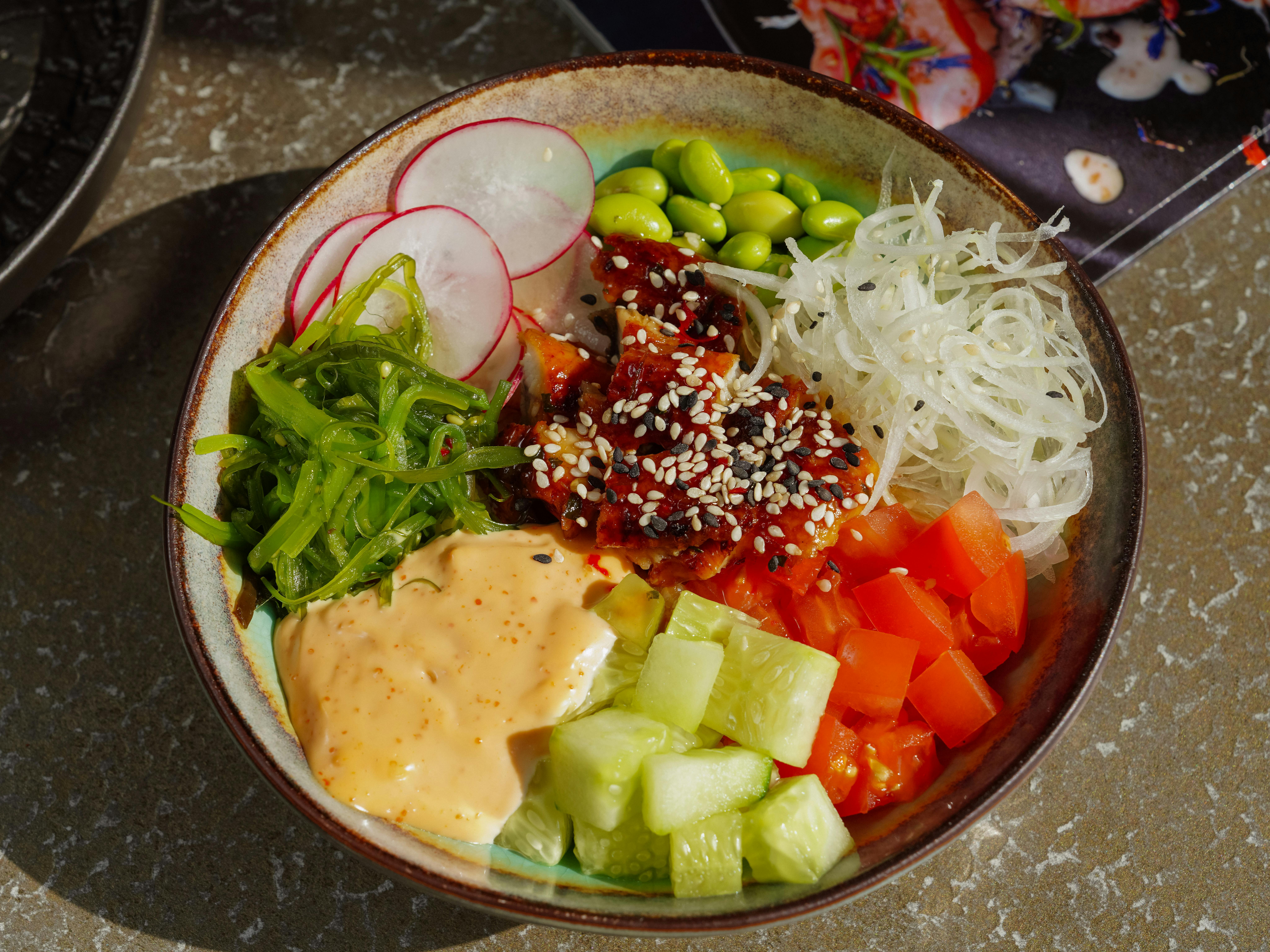 Colorful poke bowl with fresh vegetables, rice, and seafood topped with sesame seeds and sauce.